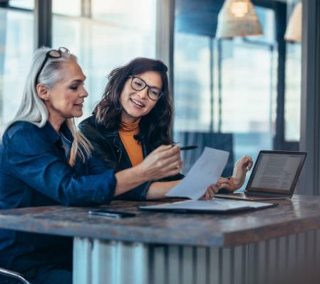 Two women analyzing documents while sitting on a table in office. Woman executives at work in office discussing some paperwork.